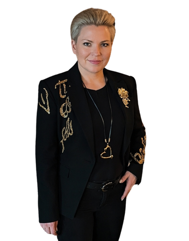 Portrait of a confident woman in a black blazer with gold embroidery, wearing a heart pendant necklace.